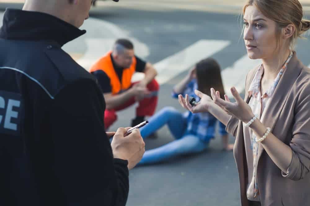 Female passenger discussing car accident with police officer, other injured passenger in background.