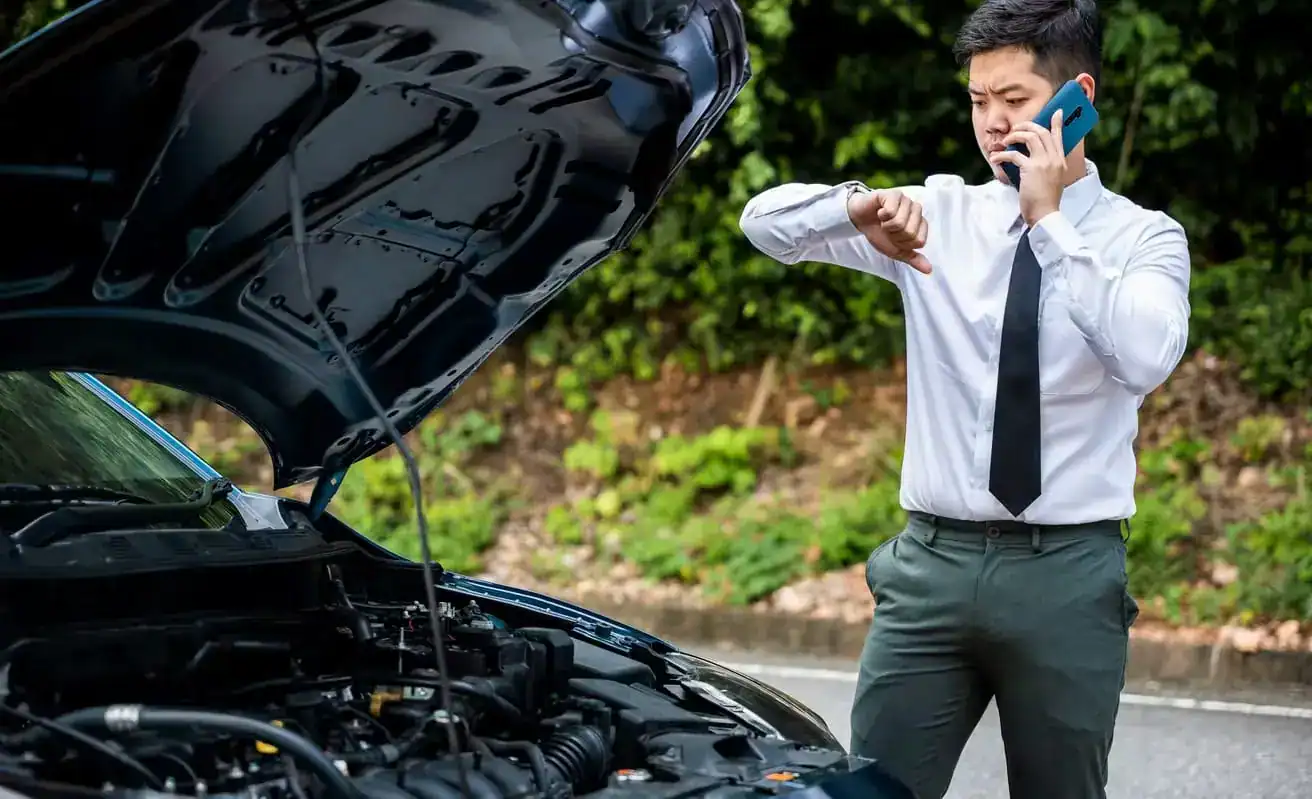 Frustrated man in business attire calling for help while stranded with a broken-down car and open hood on the roadside.