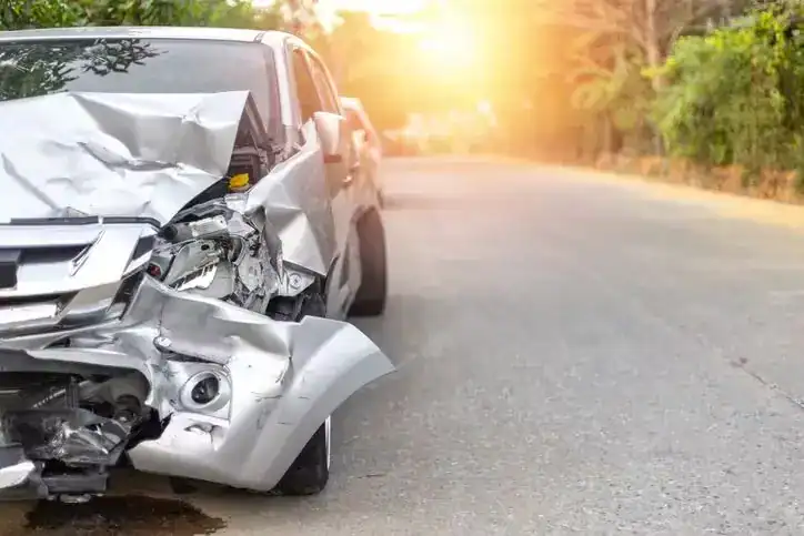 Silver car with severe front-end damage after a collision parked on the roadside at sunset.