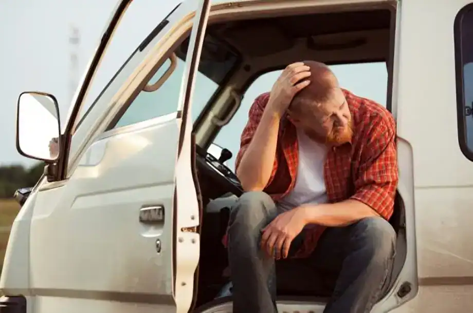 Injured truck driver sitting in cab doorway holding his head after an accident, looking distressed.