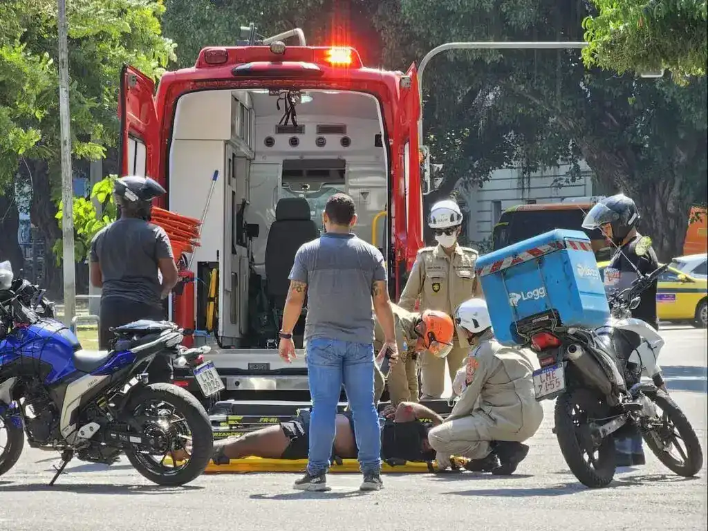 Paramedics assisting injured motorcyclist after road accident with ambulance and bystanders present.