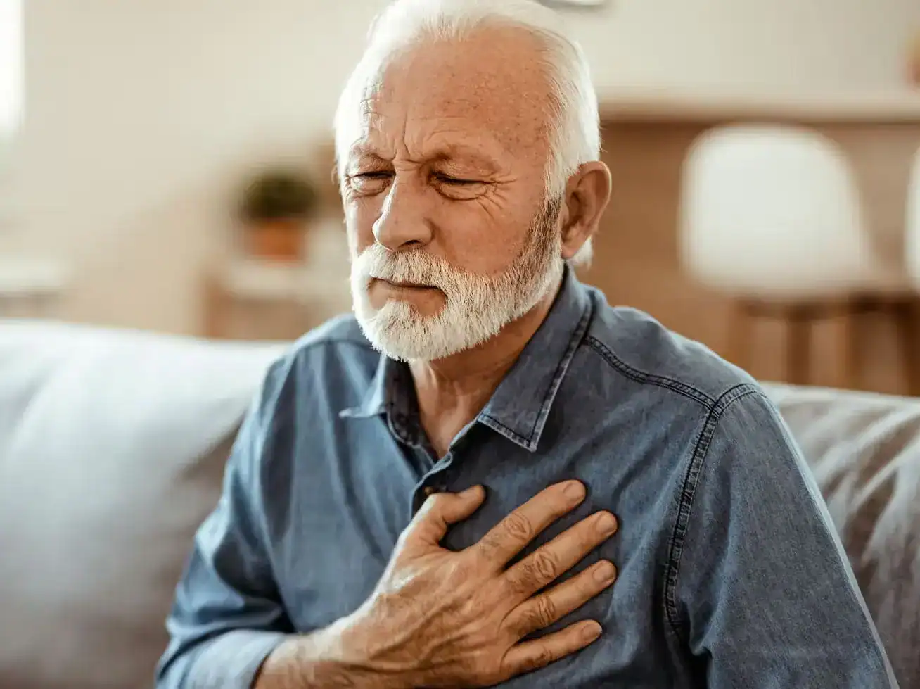 Elderly man clutching chest with pain, showing symptoms of heart attack while sitting on sofa.