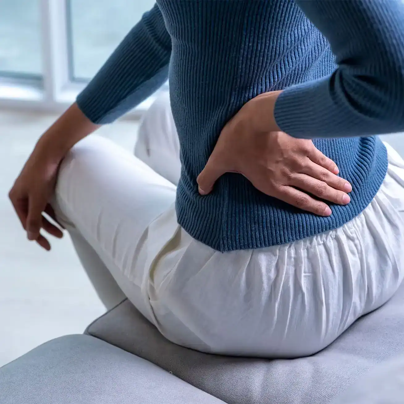 Close-up of woman holding lower back in pain from injury while sitting on couch.