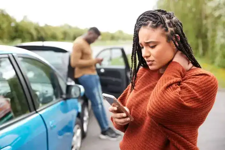 Young woman holding her neck in pain while using her phone after a car accident, with another driver standing near the vehicles.